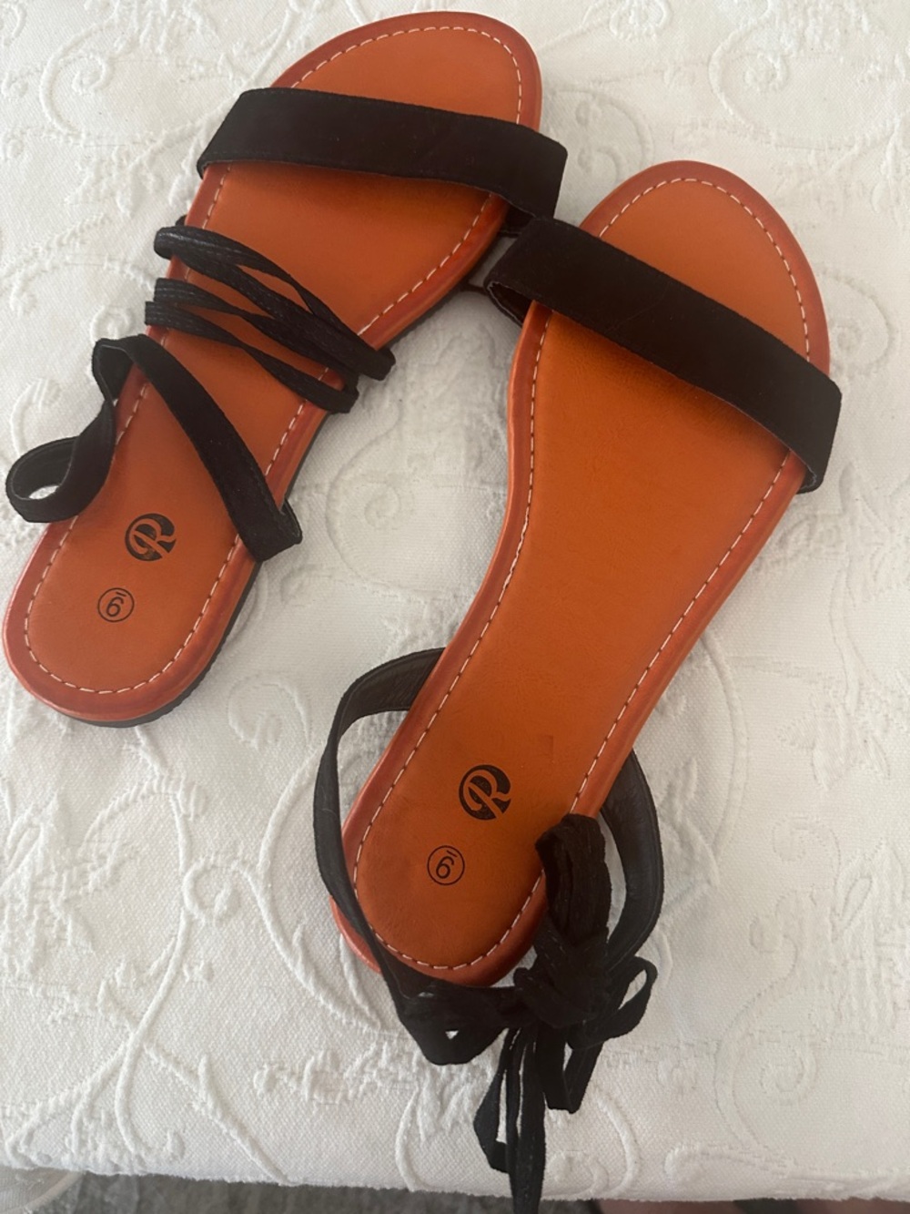 Flat sandals with a black ankle-wrap strap design and an orangish  footbed